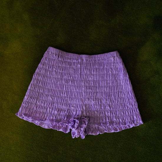 Purple smocked cotton bloomers worn with a lilac striped cardigan, showing the short silhouette and ruffled edges.